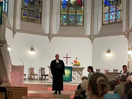 Quelle: Pfarrerin Mirjam Keim Innenraum der bunt beleuchteten Evangelischen Stadtkirche in Baden-Baden während des Aperol-Gottesdienstes am 13. September 2025. Die Besucher sitzen und schauen nach vorn, wo Dekan Christian Link in seiner schwarzen Robe vor dem Altar steht und seine Predigt hält.