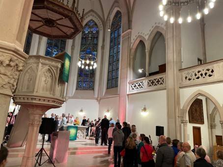 Quelle: Pfarrerin Mirjam Keim Innenraum der bunt beleuchteten Evangelischen Stadtkirche in Baden-Baden während des Aperol-Gottesdienstes am 13. September 2025. Die Gottesdienstbesucher stehen in einer Schlange, die zu dem Altar führt, neben dem sie ein Schnapsgläschen mit klassischem oder alkoholfreiem Aperol erhalten.